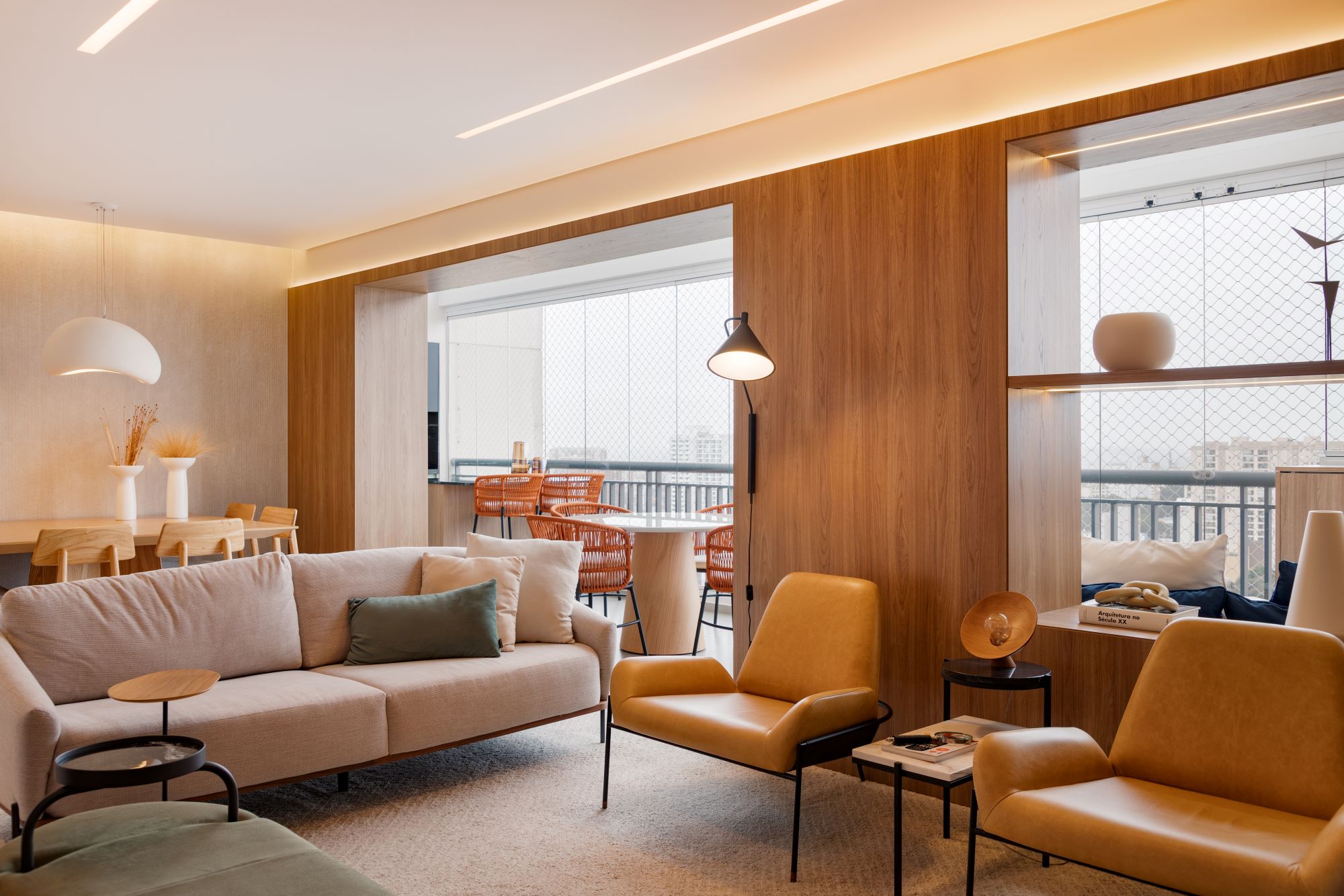Projeto FF2: sala com painel de madeira iluminado, sofá claro, duas poltronas de couro caramelo, luminária de pé preta e a varanda ao fundo